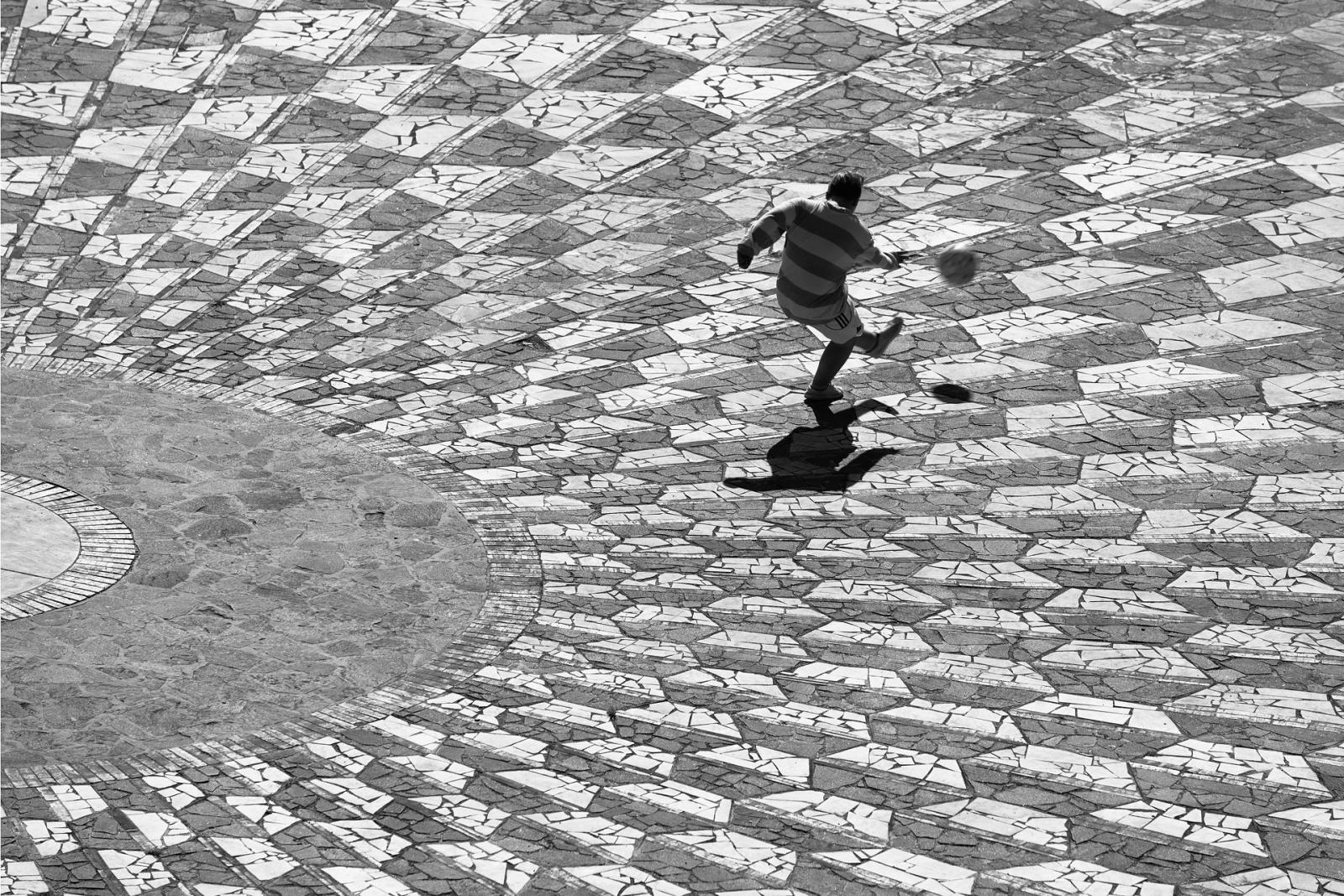 Un ragazzo calcia un pallone su una pavimentazione a mosaico, fotografia di Roberto Lugano